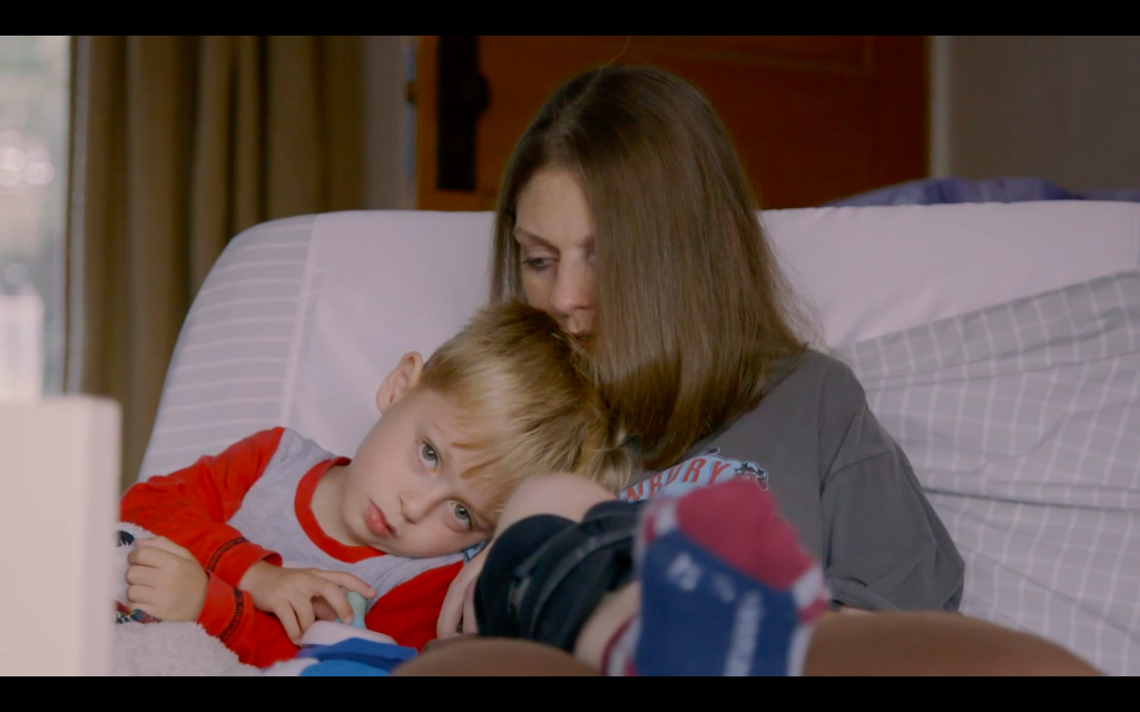 A young boy is resting on his mothers shoulder in a hospital bed.
