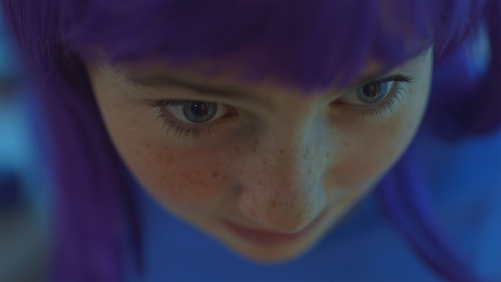 Still image from the film G_iRL. Close up photo of a girl with purple hair, freckles and blue clothes. 